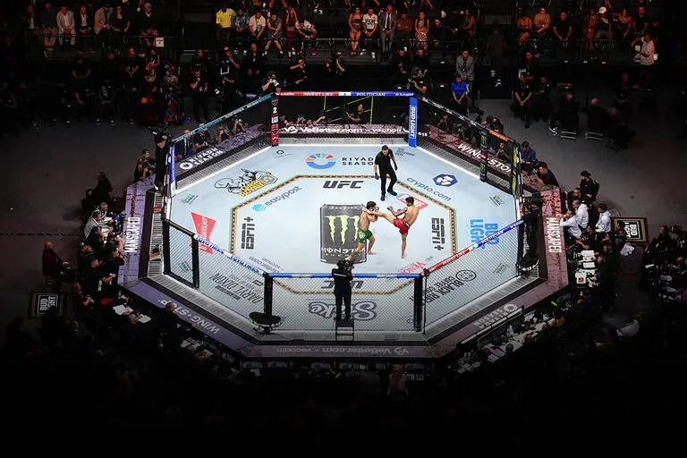 The octagon from above during a live event: two fighters mid-exchange in the centre of the cage, the referee circling, and cornermen, camera operators and the crowd ringing it on every side.